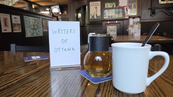 A "Writers of Ottawa" sign sits on a wooden table next to a glass bulb of tea and a white tea mug. In the background is the wood-paneled wall of the pub covered in signs and framed art.
